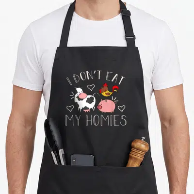Homy vegan apron - I DON'T EAT MY HOMIES text over cartoon cow pig rooster and hearts — shown on a black cotton canvas apron against a white background