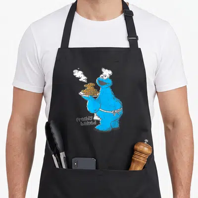 Freshly cookie monster apron - Blue character holding stack of orange cookies with steam and text Freshly baked — shown on a black cotton canvas apron against a white background