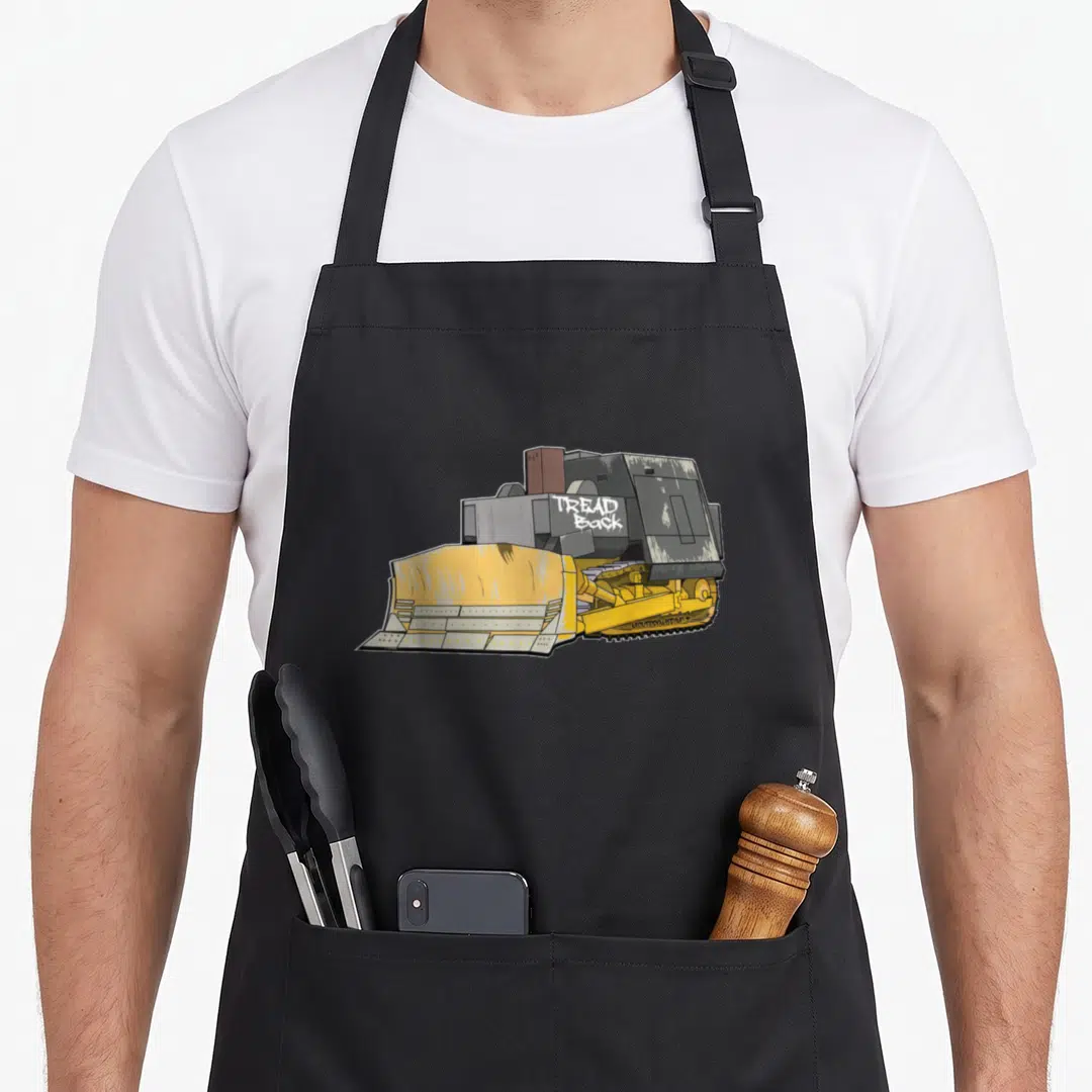 Killdozer bulldozer girl apron - Cartoon bulldozer in yellow and grey with white TREAD BACK graffiti text on side — shown on a black cotton canvas apron against a white background