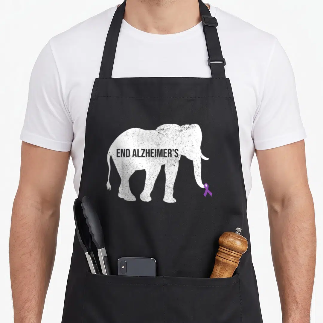 End alzheimer's aprons - Distressed white elephant silhouette holding purple ribbon with text END ALZHEIMER'S across — shown on a black cotton canvas apron against a white background
