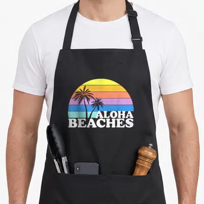Aloha tropical apron - Retro palm trees in front of striped sun with ALOHA BEACHES text at bottom right — shown on a black cotton canvas apron against a white background