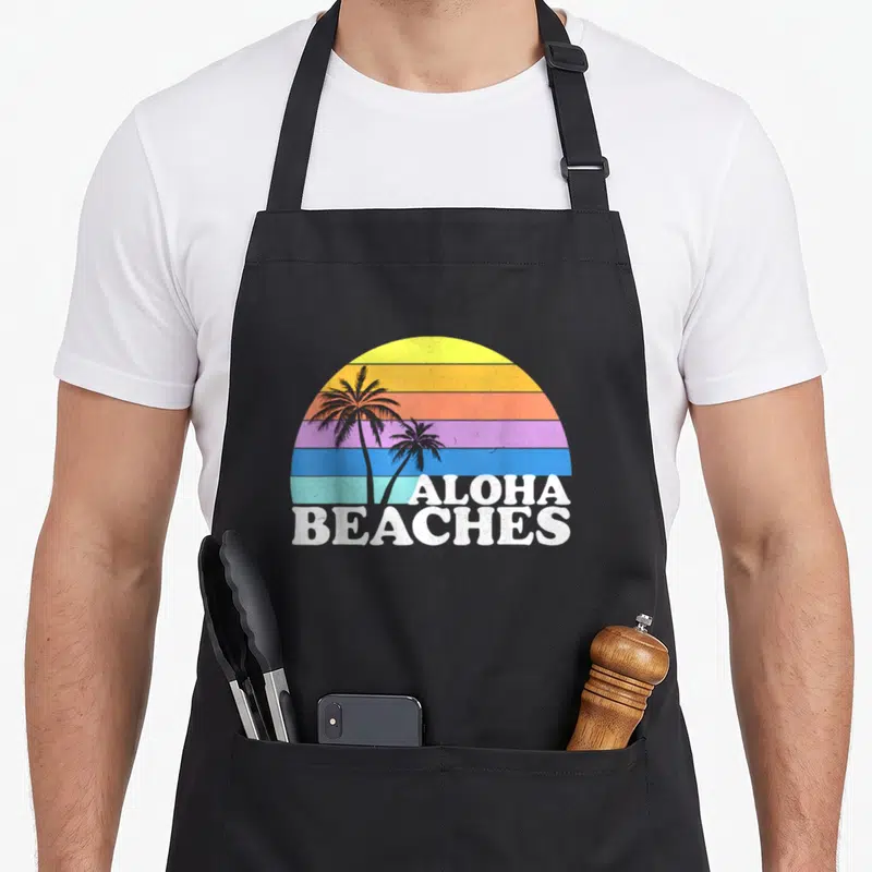 Aloha tropical apron - Retro palm trees in front of striped sun with ALOHA BEACHES text at bottom right — shown on a black cotton canvas apron against a white background