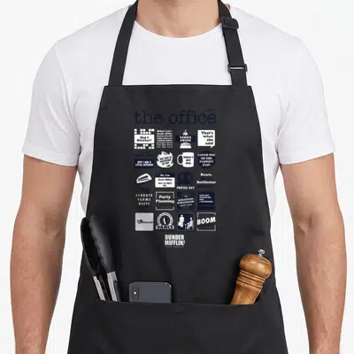 Nbc x apron - Grid of The Office quotes and logos including Did I Stutter and World's Best Boss in blue and black — shown on a black cotton canvas apron against a white background