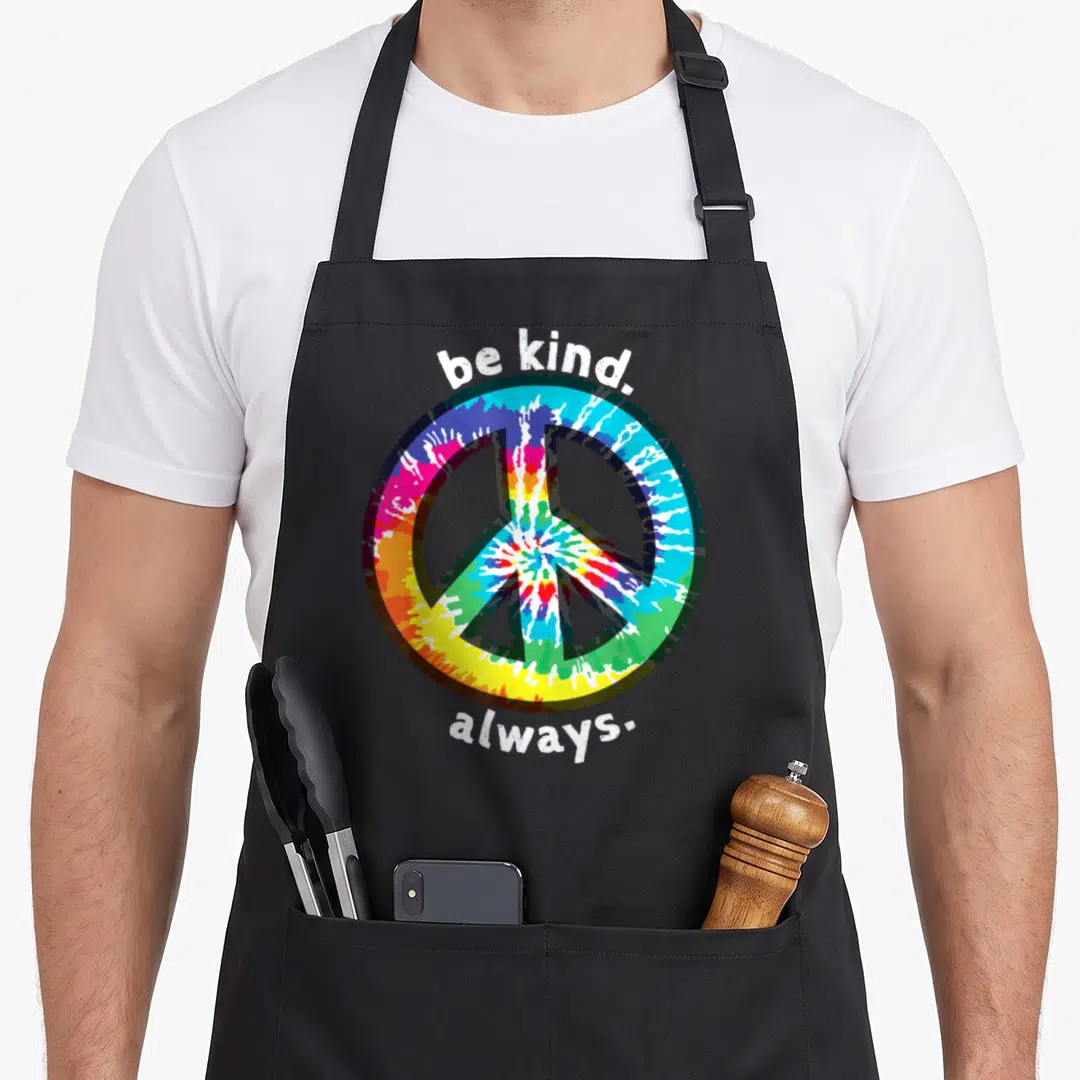 Kind tie-dye apron - Tie-dye peace symbol with be kind always text in white on black fabric — shown on a black cotton canvas apron against a white background