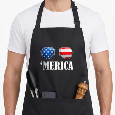 Independence patriot 4th of july apron - Sunglasses with flag lenses and MERICA text below — shown on a black cotton canvas apron against a white background