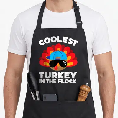Toddler thanksgiving apron - Cartoon turkey with sunglasses and blue cap reading COOLEST TURKEY IN THE FLOCK — shown on a black cotton canvas apron against a white background