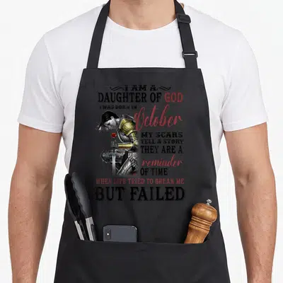 October the story of the apron - Woman warrior in gold armor holding sword with red script text I Am A Daughter Of God — shown on a black cotton canvas apron against a white background