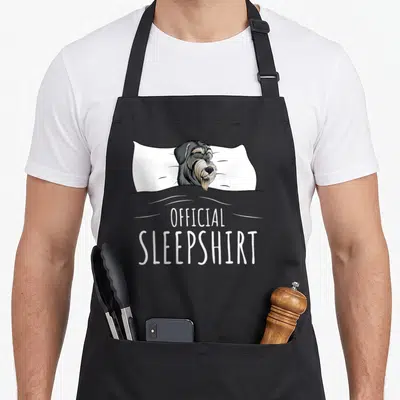 Pajama miniature schnauzer apron - Sleeping miniature schnauzer on pillow with official sleepshirt text between two — shown on a black cotton canvas apron against a white background
