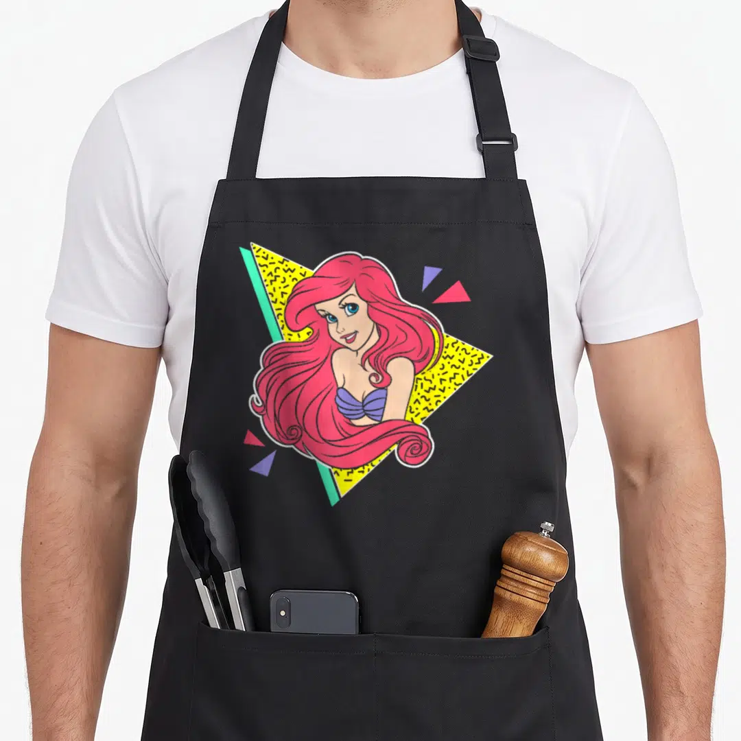 80s mermaid apron - Cartoon woman with pink hair over yellow and teal triangles with purple accents — shown on a black cotton canvas apron against a white background