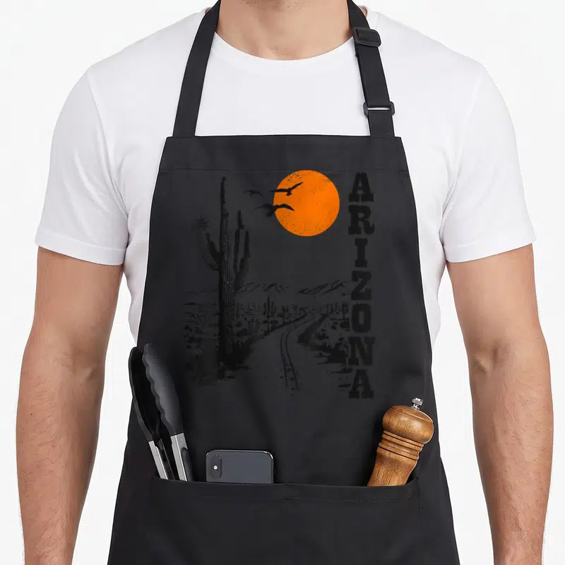 Arizona western apron - Retro black and orange desert print with vertical ARIZONA text, cactus, sun, and flying birds — shown on a black cotton canvas apron against a white background