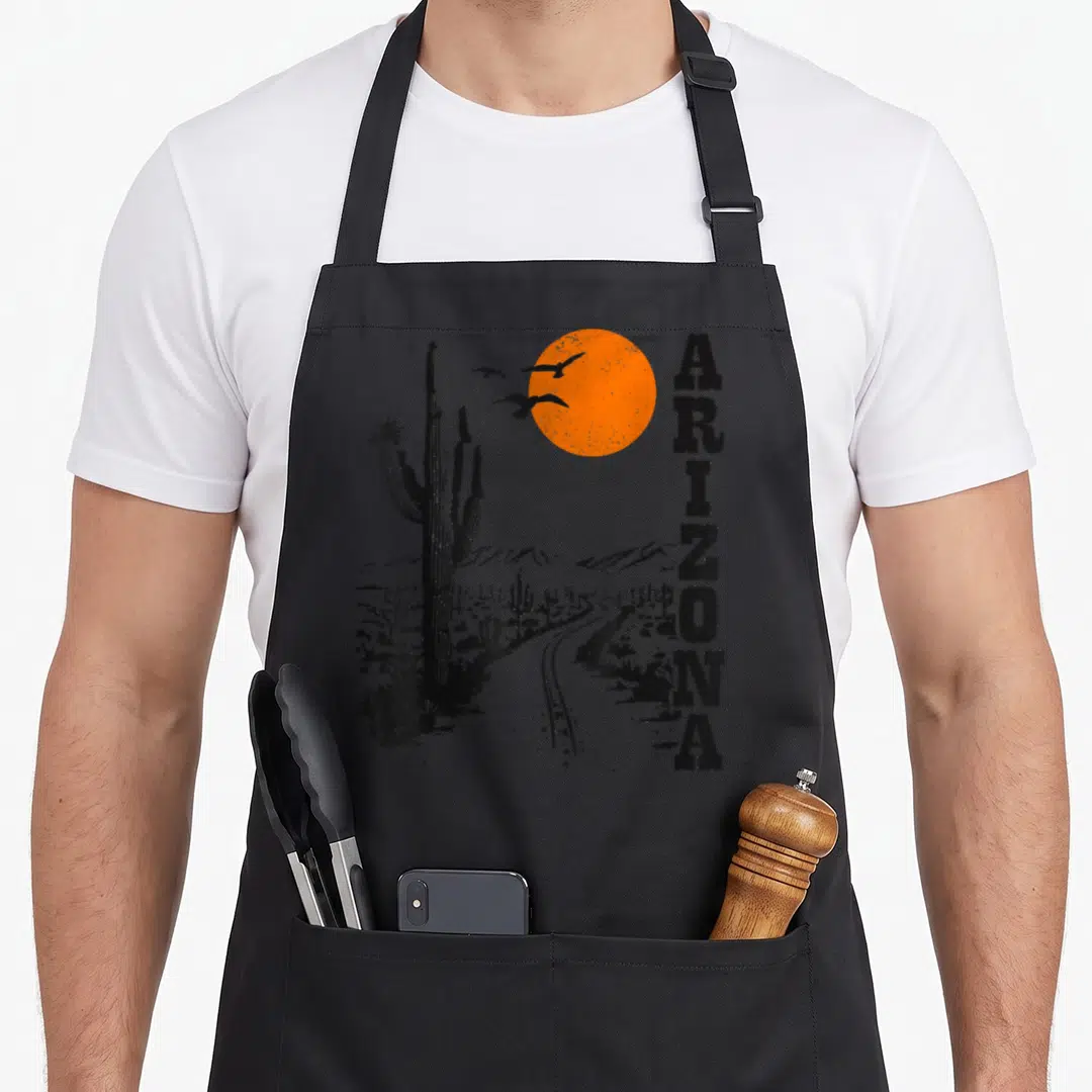 Arizona western apron - Retro black and orange desert print with vertical ARIZONA text, cactus, sun, and flying birds — shown on a black cotton canvas apron against a white background