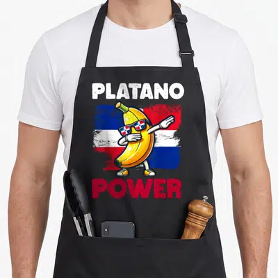 Platano dominican apron - Dabbing banana wearing flag sunglasses with PLATANO POWER text — shown on a black cotton canvas apron against a white background