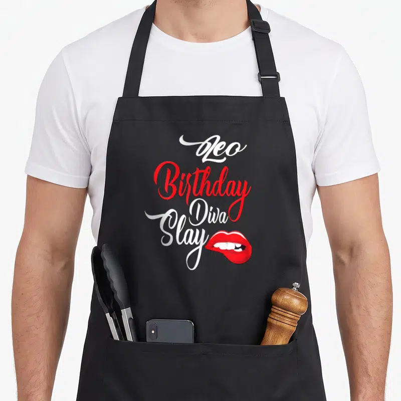Bite birthday apron - Stacked Leo Birthday Diva Slay text with red lips graphic — shown on a black cotton canvas apron against a white background