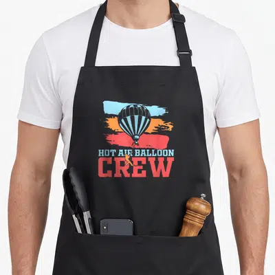 Crew balloon apron - Retro hot air balloon with small figure on rope over blue, orange, and red brush strokes — shown on a black cotton canvas apron against a white background