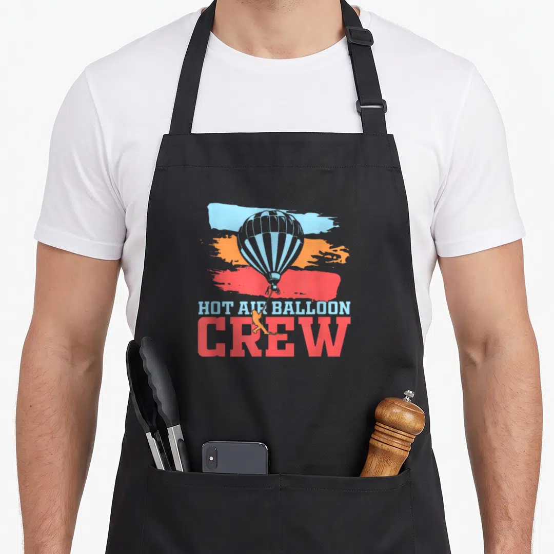 Crew balloon apron - Retro hot air balloon with small figure on rope over blue, orange, and red brush strokes — shown on a black cotton canvas apron against a white background