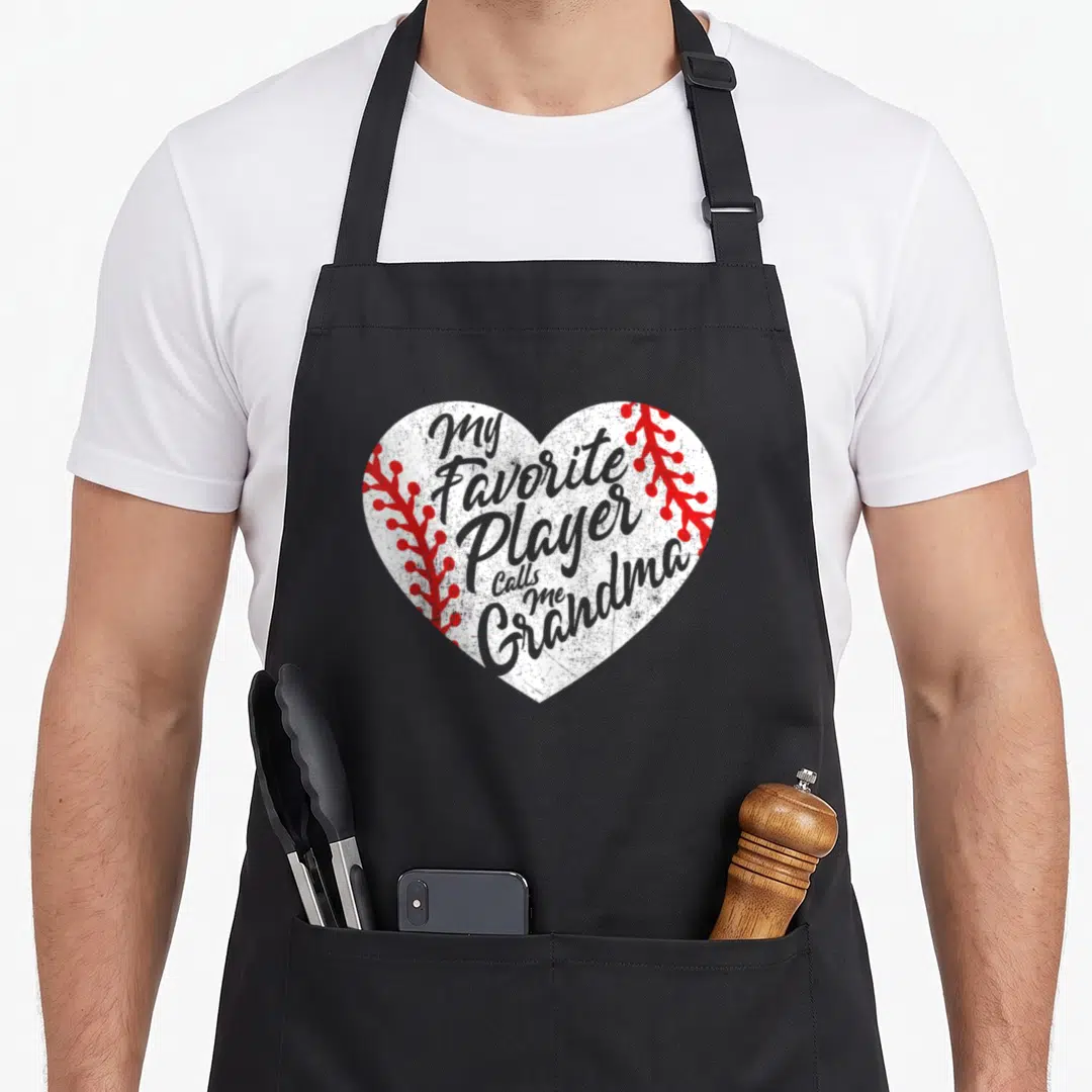 Player grandma apron - White heart with red stitching containing grey text My Favorite Player Calls Me Grandma — shown on a black cotton canvas apron against a white background