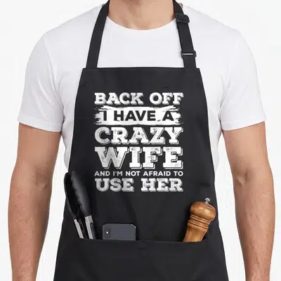 Afraid wife apron - White stacked typography reading BACK OFF I HAVE A CRAZY WIFE AND I M NOT AFRAID TO USE — shown on a black cotton canvas apron against a white background