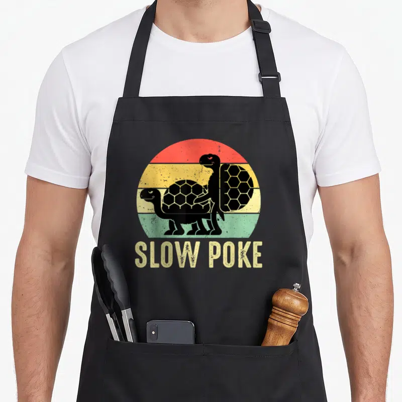 Slow poke apron - Two black turtles over red yellow green striped sun with SLOW POKE text below — shown on a black cotton canvas apron against a white background
