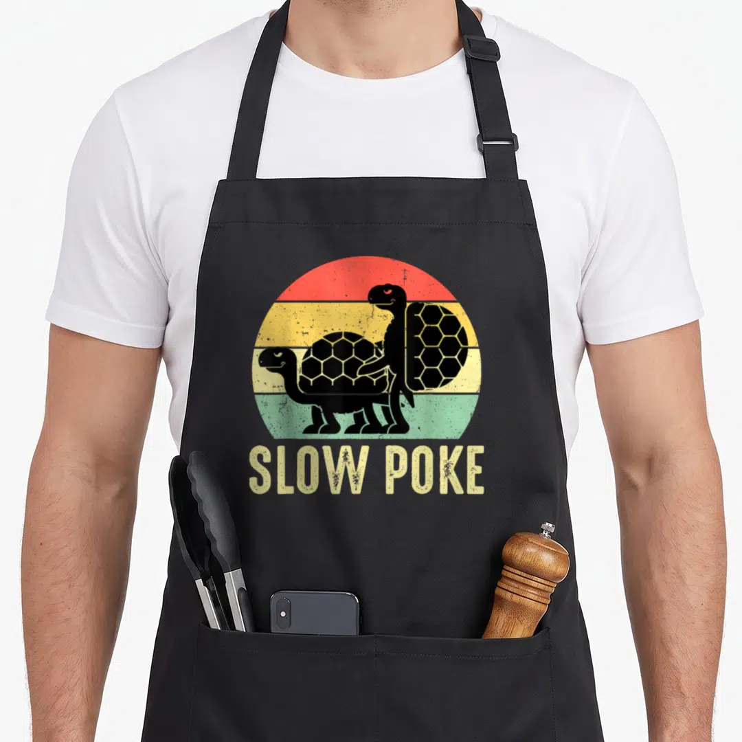 Slow poke apron - Two black turtles over red yellow green striped sun with SLOW POKE text below — shown on a black cotton canvas apron against a white background