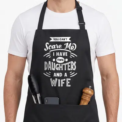 Scare daughter wife apron - White stacked text reading YOU CAN'T Scare Me I HAVE TWO DAUGHTERS AND A WIFE with banner — shown on a black cotton canvas apron against a white background
