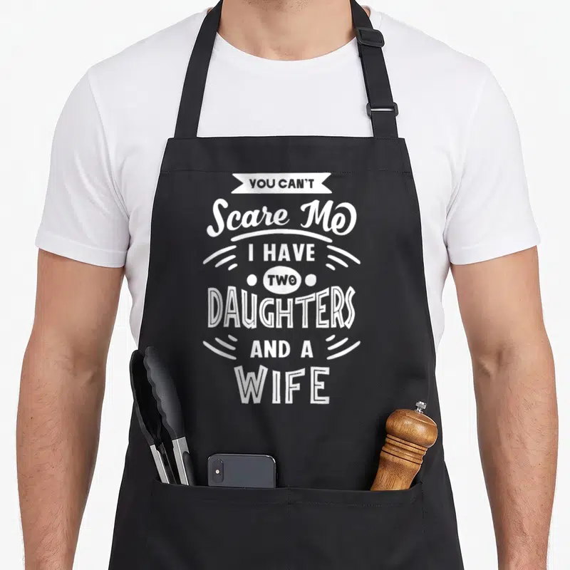 Scare daughter wife apron - White stacked text reading YOU CAN'T Scare Me I HAVE TWO DAUGHTERS AND A WIFE with banner — shown on a black cotton canvas apron against a white background