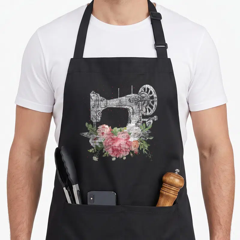Sewing craft apron - Distressed white sewing machine with pink and green watercolor flower cluster overlay — shown on a black cotton canvas apron against a white background