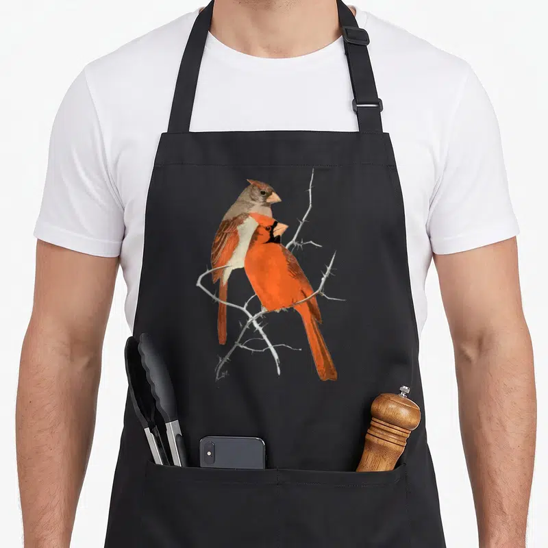 Funny bird cardinal apron - Two realistic cardinals in red and brown perched on a thorny grey branch — shown on a black cotton canvas apron against a white background