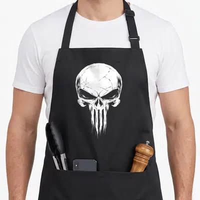Punisher distressed apron - White stylized skull with cracked forehead and dripping jaw printed on grey background — shown on a black cotton canvas apron against a white background