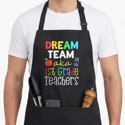 Dream aka teacher apron - Stacked colorful text reading Dream Team aka 1st Grade Teachers with apple and two crayons — shown on a black cotton canvas apron against a white background