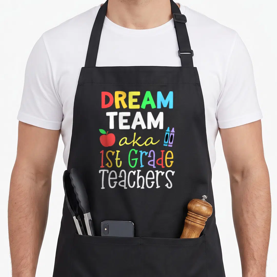 Dream aka teacher apron - Stacked colorful text reading Dream Team aka 1st Grade Teachers with apple and two crayons — shown on a black cotton canvas apron against a white background