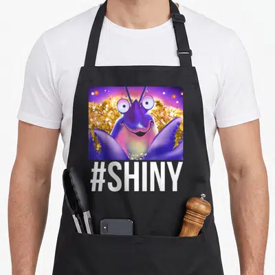 Shiny novelty apron - Cartoon purple insect in gold glitter square with #SHINY text below — shown on a black cotton canvas apron against a white background