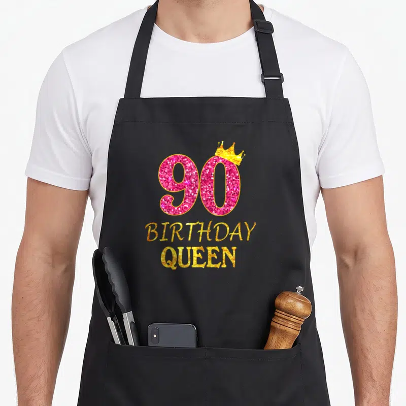 90th queen apron - Pink glitter number 90 with gold crown and stacked birthday queen text — shown on a black cotton canvas apron against a white background