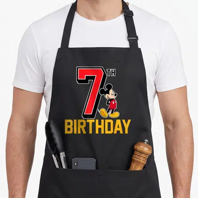 Funny 7th birthday apron - Red number 7 with leaning cartoon character and yellow BIRTHDAY text — shown on a black cotton canvas apron against a white background