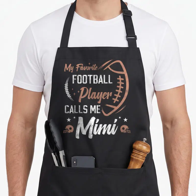 Mimi grandma aprons - Distressed text My Favorite Football Player Calls Me Mimi with orange football outline and helmets — shown on a black cotton canvas apron against a white background