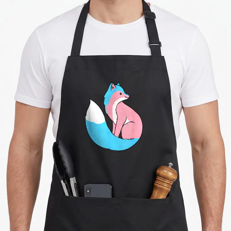Tran fox print apron - Cartoon fox in blue pink and white transgender pride colors — shown on a black cotton canvas apron against a white background