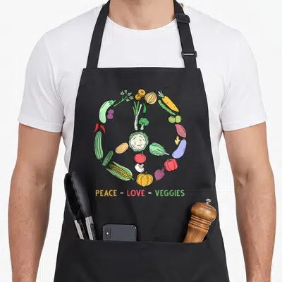 Themed veggie apron - Vegetables arranged in a circle forming a peace sign with text PEACE - LOVE - VEGGIES below — shown on a black cotton canvas apron against a white background
