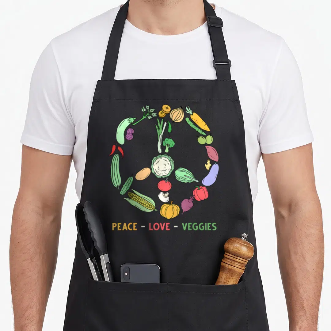 Themed veggie apron - Vegetables arranged in a circle forming a peace sign with text PEACE - LOVE - VEGGIES below — shown on a black cotton canvas apron against a white background