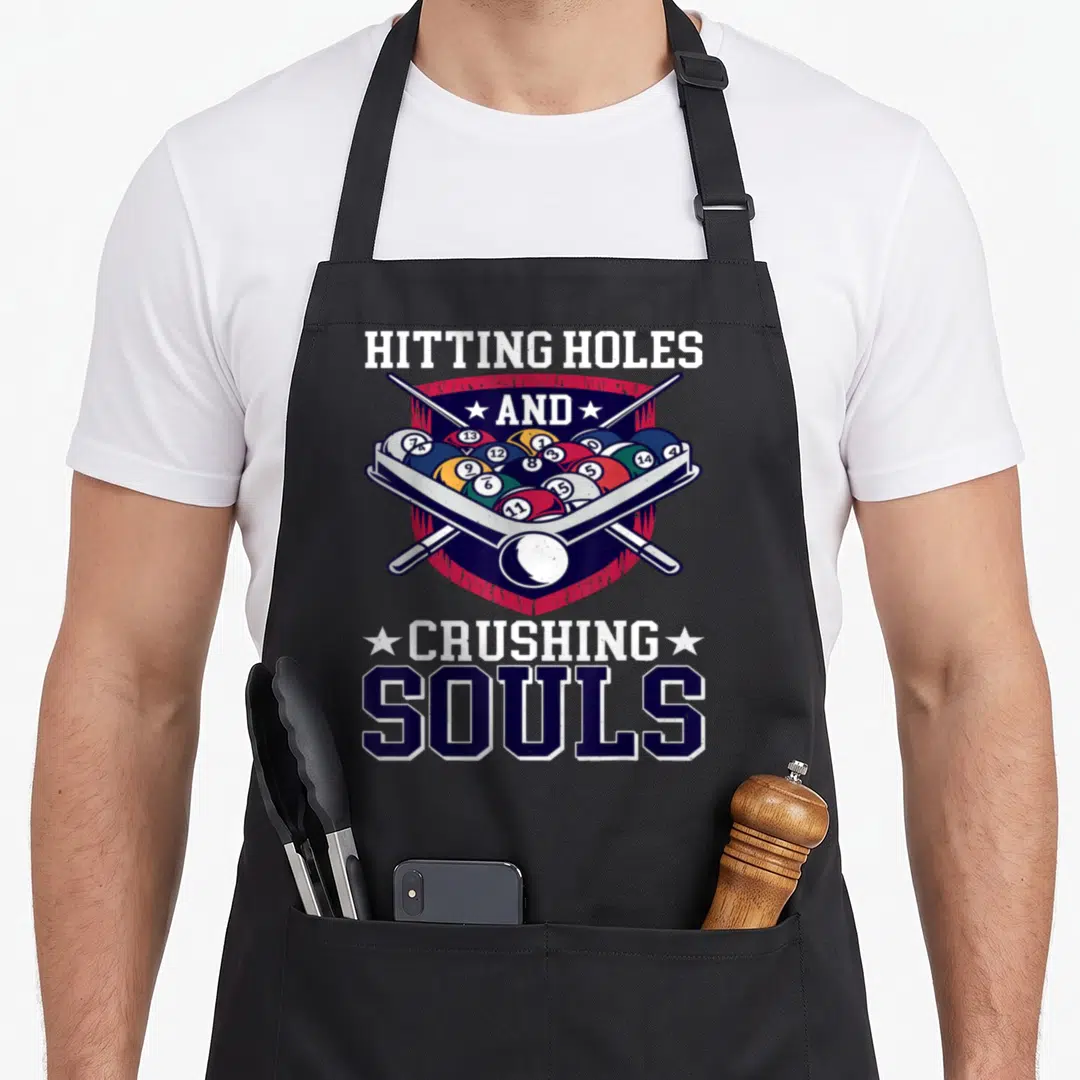 Accessory billiards apron - HITTING HOLES AND CRUSHING SOULS text above billiard balls on a shield with crossed cues — shown on a black cotton canvas apron against a white background