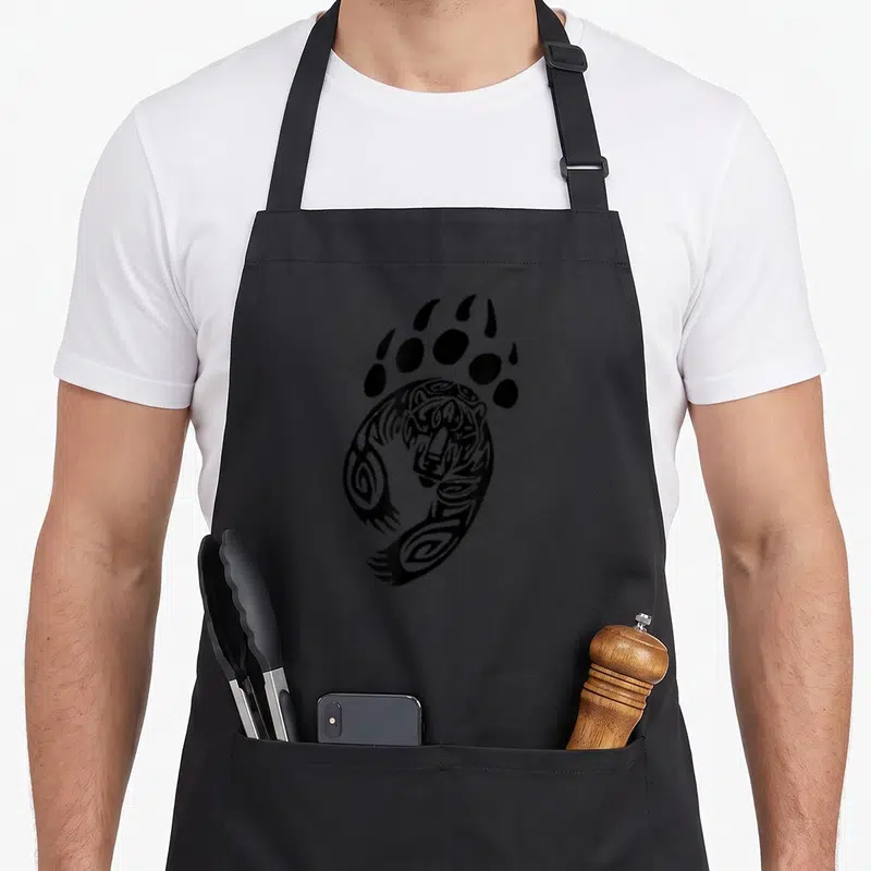 Paw bear apron - Black tribal line art bear head inside a large paw print footprint design — shown on a black cotton canvas apron against a white background
