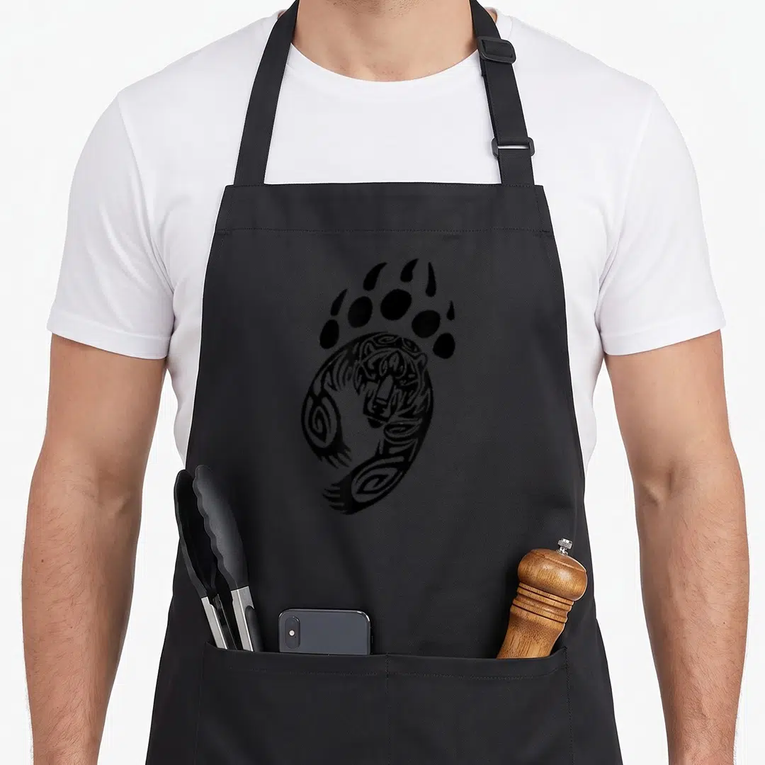 Paw bear apron - Black tribal line art bear head inside a large paw print footprint design — shown on a black cotton canvas apron against a white background
