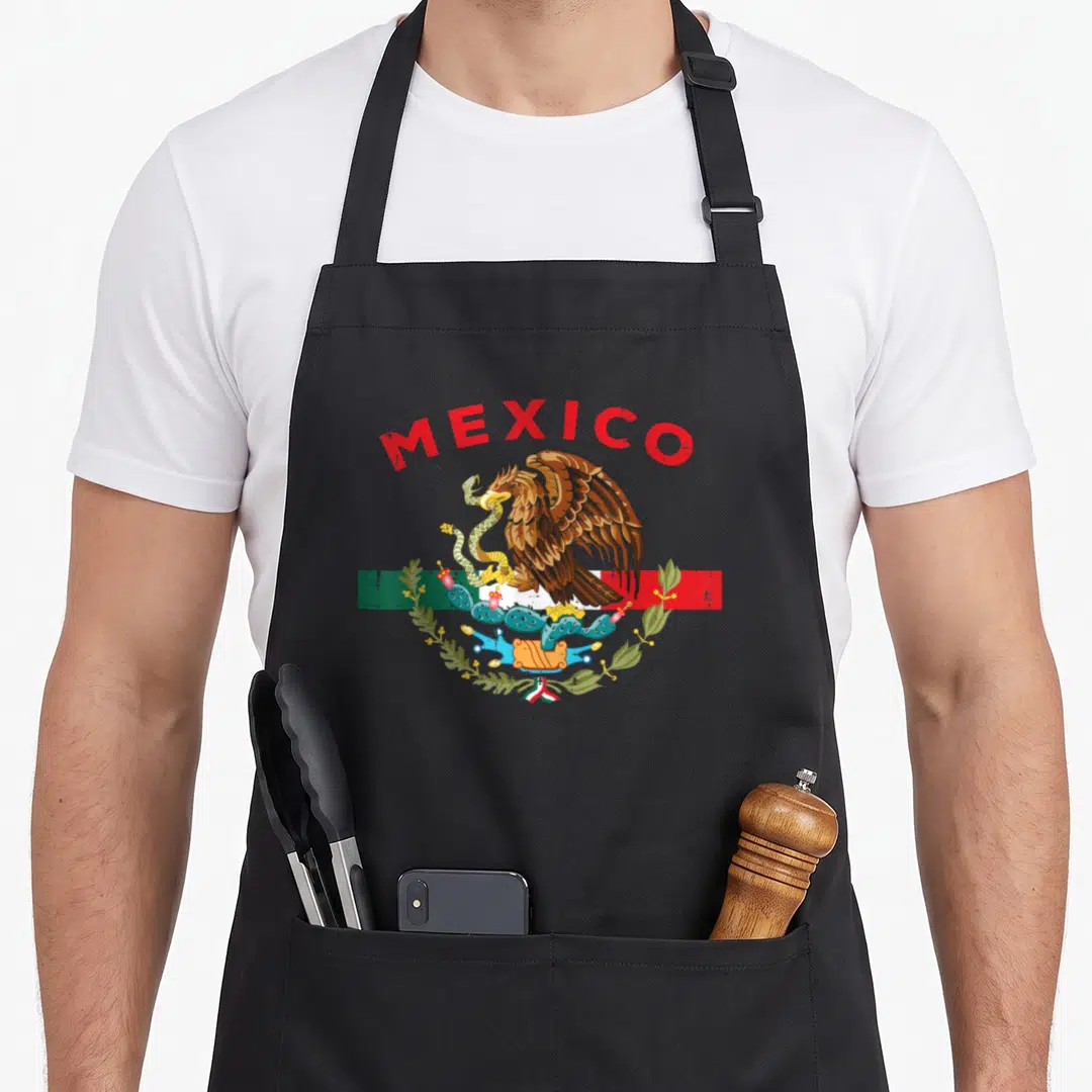 Eagle mexican apron - MEXICO text arching over an eagle perched on a cactus eating a snake against a flag background — shown on a black cotton canvas apron against a white background