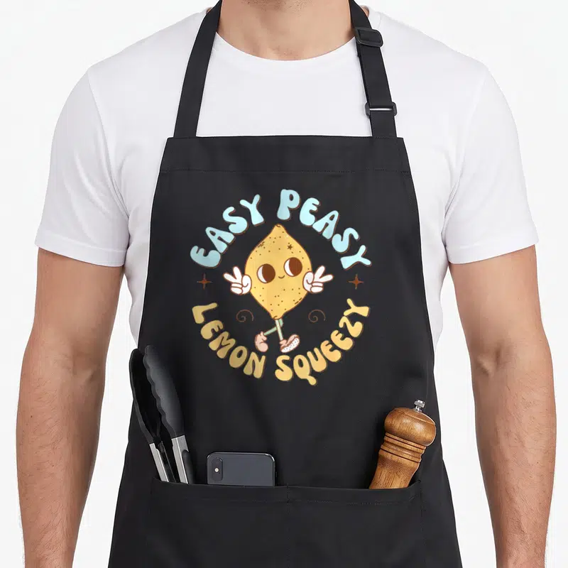 Peasy lemon apron - Retro cartoon lemon character with peace signs and arched text EASY PEASY LEMON SQUEEZY in yellow — shown on a black cotton canvas apron against a white background