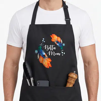 Pet betta fish apron - Two watercolor betta fish in orange and blue curving around white Betta Mom script text — shown on a black cotton canvas apron against a white background