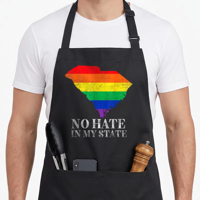 Hate gay pride apron - Rainbow South Carolina map with distressed white text reading NO HATE IN MY STATE — shown on a black cotton canvas apron against a white background