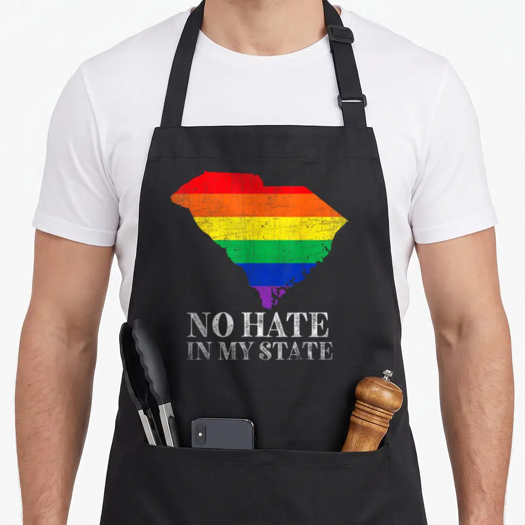 Hate gay pride apron - Rainbow South Carolina map with distressed white text reading NO HATE IN MY STATE — shown on a black cotton canvas apron against a white background