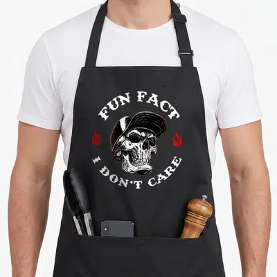 Fact skeleton apron - Realistic skull in cap with Fun Fact I Don't Care text and red flames — shown on a black cotton canvas apron against a white background