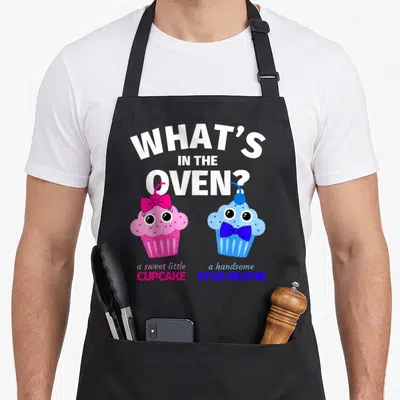 Gender muffin man apron - Arched text What's In The Oven above pink cupcake and blue muffin cartoons — shown on a black cotton canvas apron against a white background