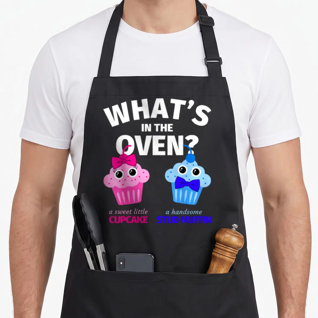 Gender muffin man apron - Arched text What's In The Oven above pink cupcake and blue muffin cartoons — shown on a black cotton canvas apron against a white background