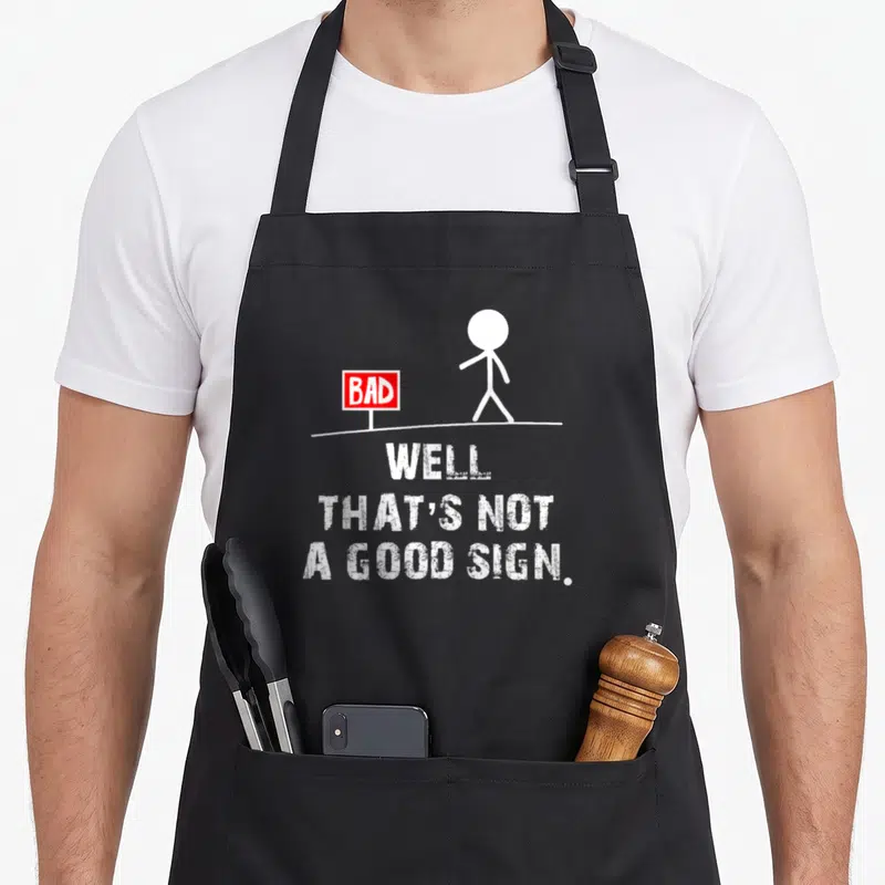 Well sarcastic apron - Stick figure walks past red bad sign above white text well that's not a good sign — shown on a black cotton canvas apron against a white background