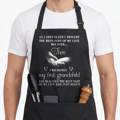 Handed grandma apron - White text and photo of hands holding on grey background with two hearts — shown on a black cotton canvas apron against a white background